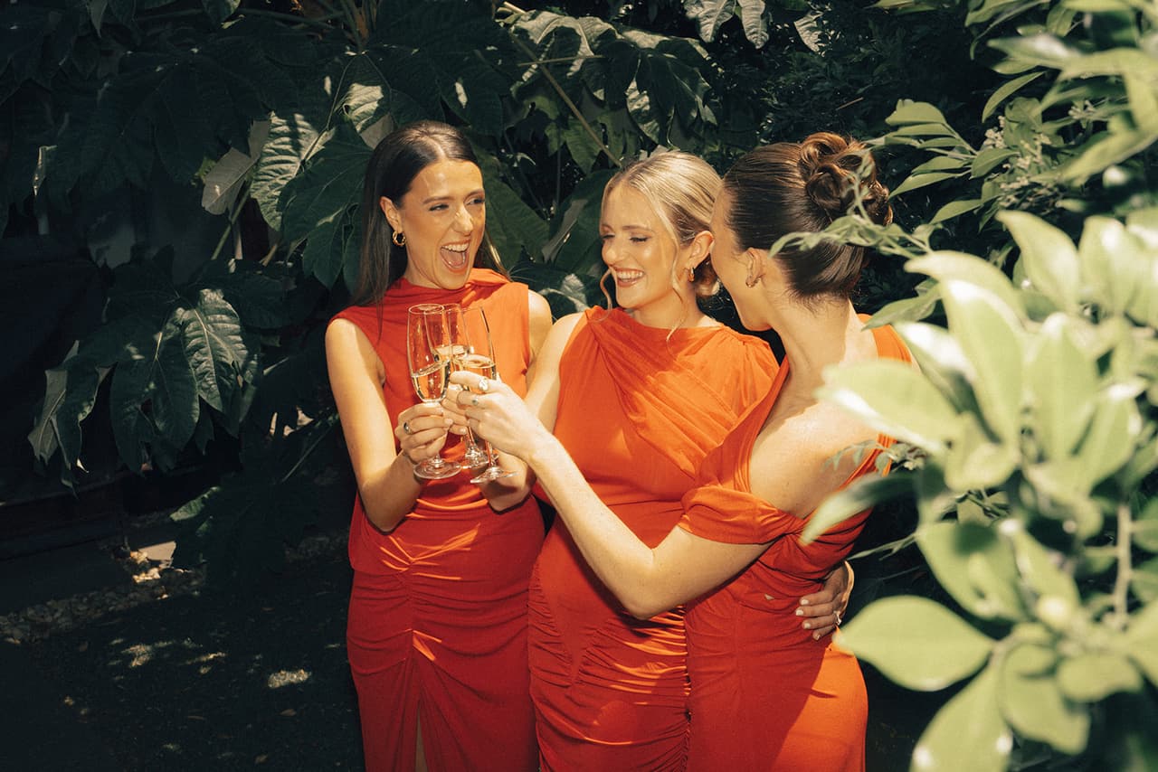 Three bridesmaids in red dresses clinking glasses and laughing under the trees