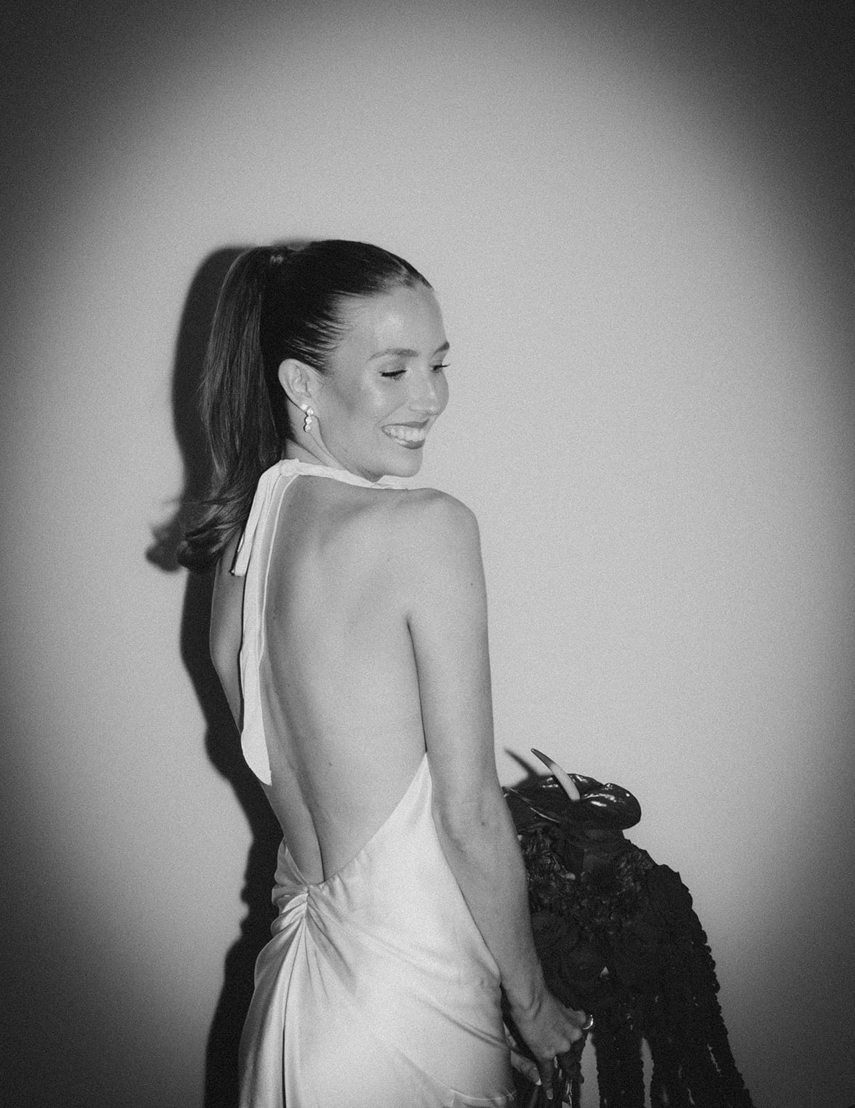 Black and white photo of Kira looking over her shoulder in a backless satin dress, her bouquet at her side