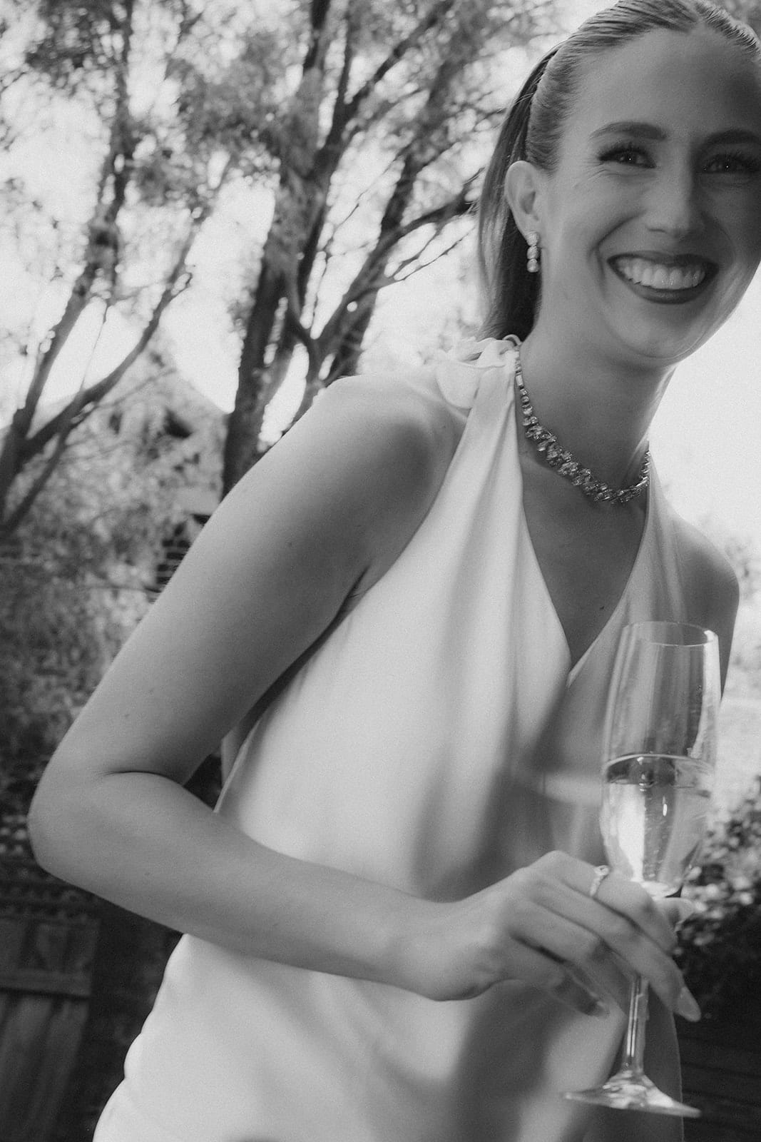 Black and white photo of Kira laughing in a white halter dress, a glass of champagne in her hand