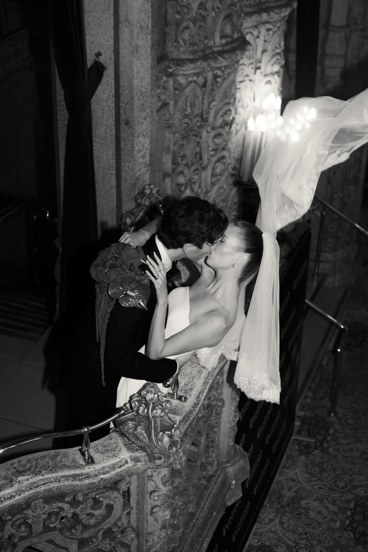 Black and white photo from above of Nick and Kira kissing on a carved staircase, her veil lifting over the balustrade