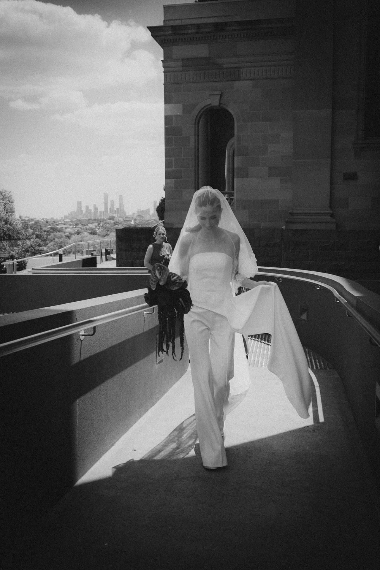 Black and white photo of Kira in a strapless jumpsuit and long veil walking along a stone terrace, the city skyline behind her