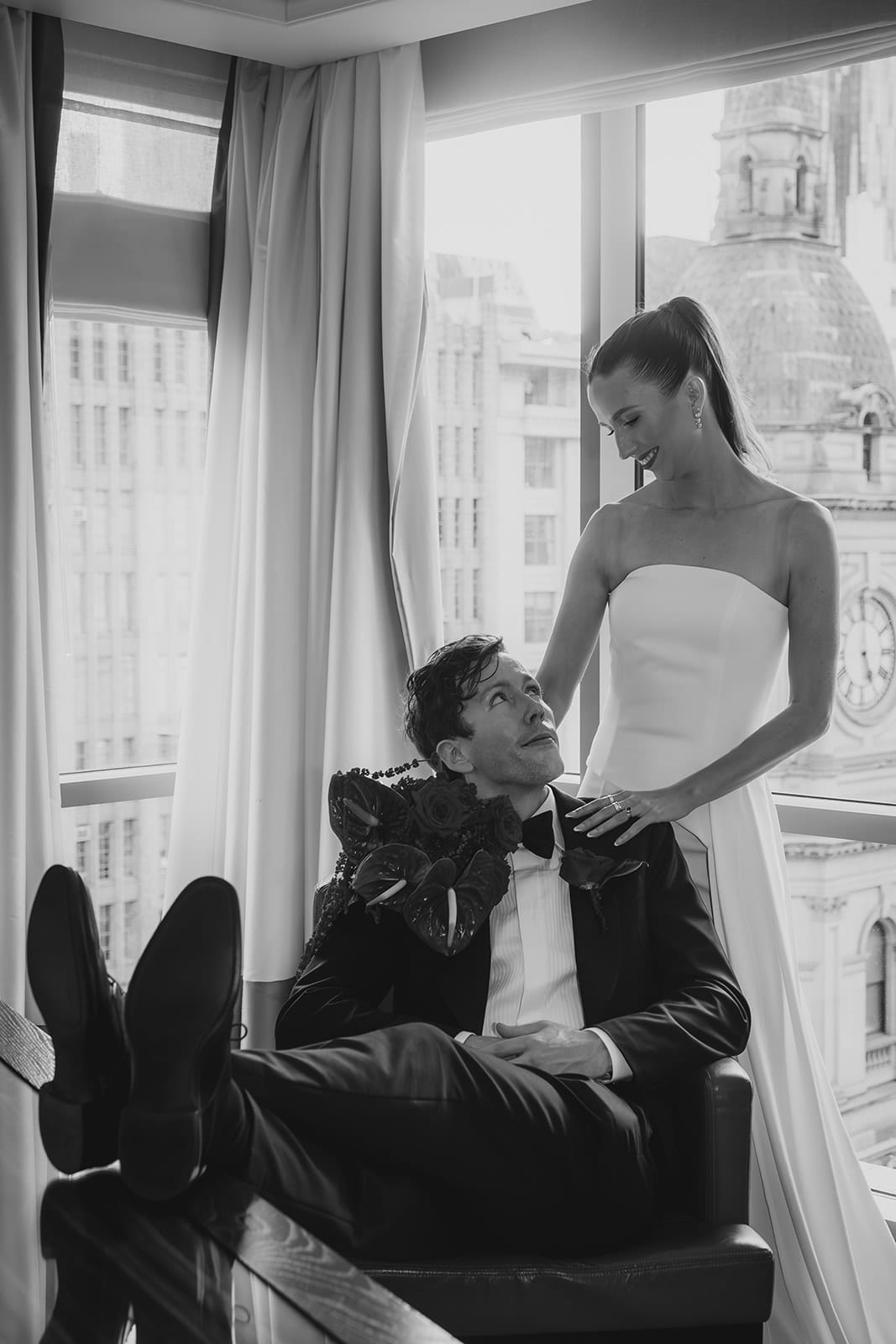 Black and white photo of Nick lounging with his feet up at the hotel window while Kira stands beside him