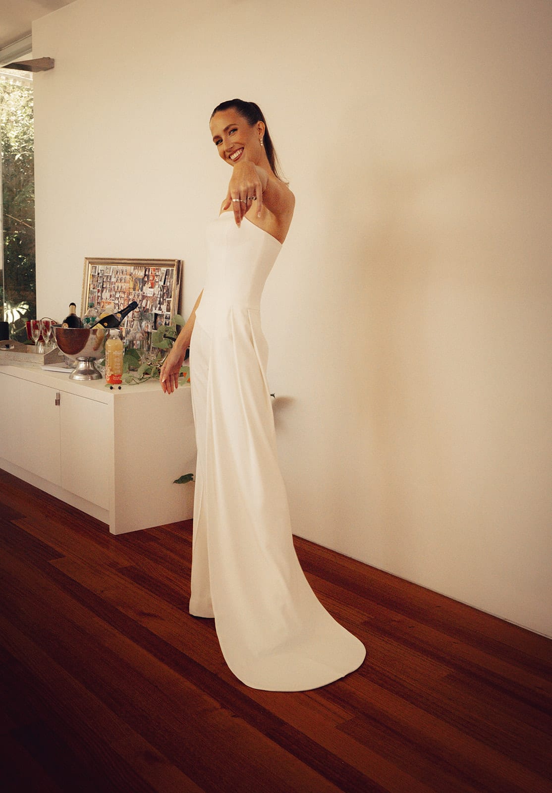 Kira in a strapless white jumpsuit with a long train, standing in a white room at home