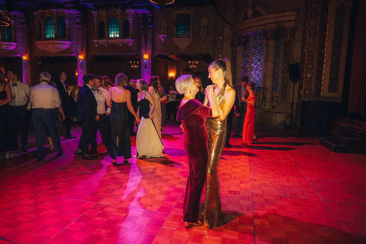 Kira in a gold sequined jumpsuit dancing with an older woman in burgundy velvet under pink light