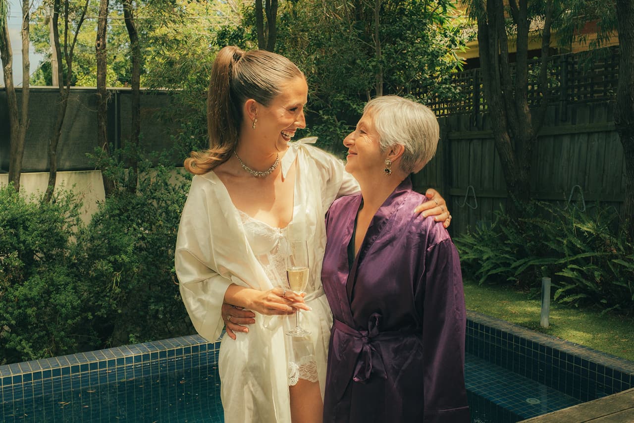 Kira in a white robe laughing with an older woman in a purple robe beside the pool