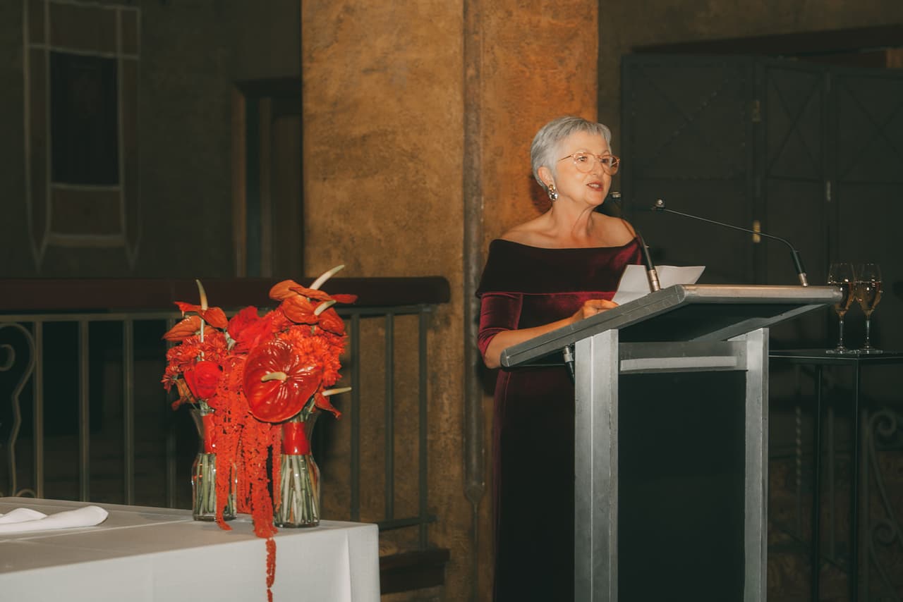 An older woman in burgundy velvet giving a speech at the lectern beside a vase of red anthuriums