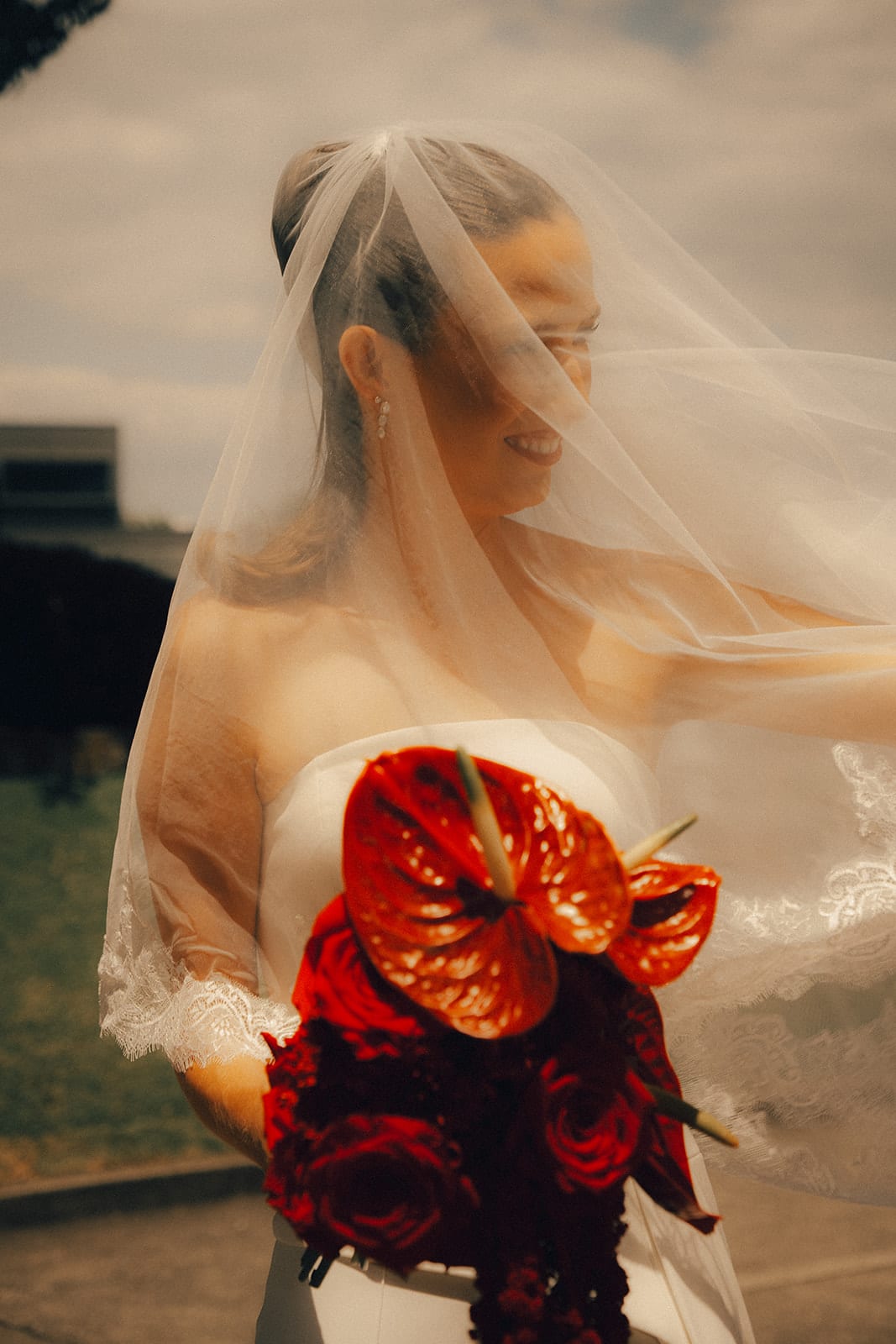Kira’s veil lifting in the wind as she holds her bouquet of red anthuriums and roses outside the chapel
