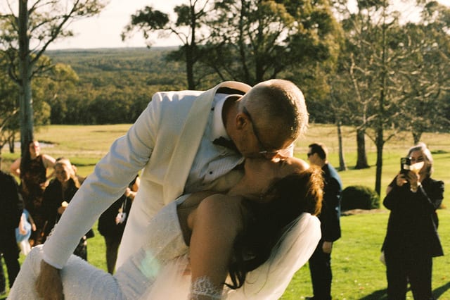 Gareth dipping Kristie for a kiss as guests take photos behind them, on 35mm film