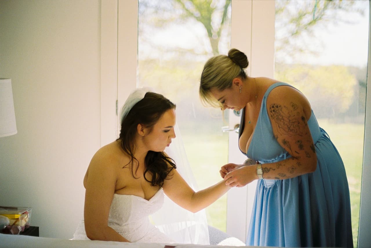 A bridesmaid helping Kristie get ready by a bright window, on 35mm film