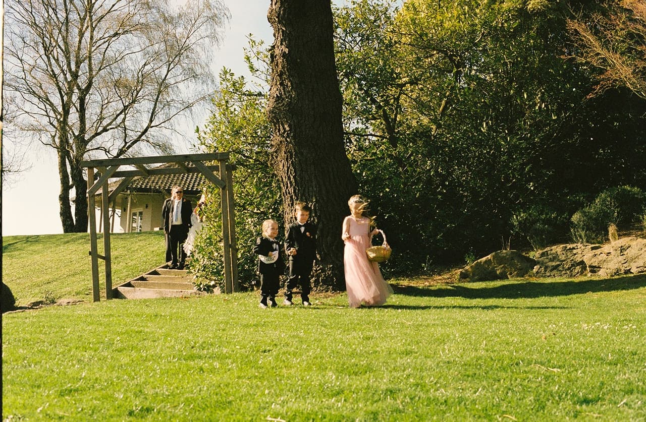 Two page boys in little suits and a flower girl in pink coming down the lawn from the timber pergola, on 35mm film