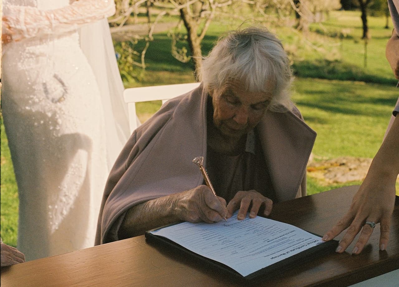 An older woman in a pink shawl signing the marriage certificate as a witness while Kristie holds the page, on 35mm film
