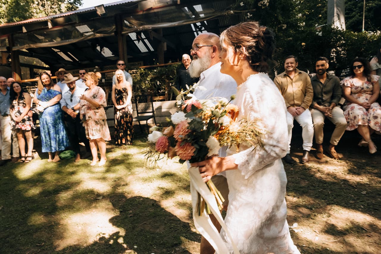 Lana walking down the aisle on her dad’s arm, her bouquet of dried flowers and ribbons in her hand