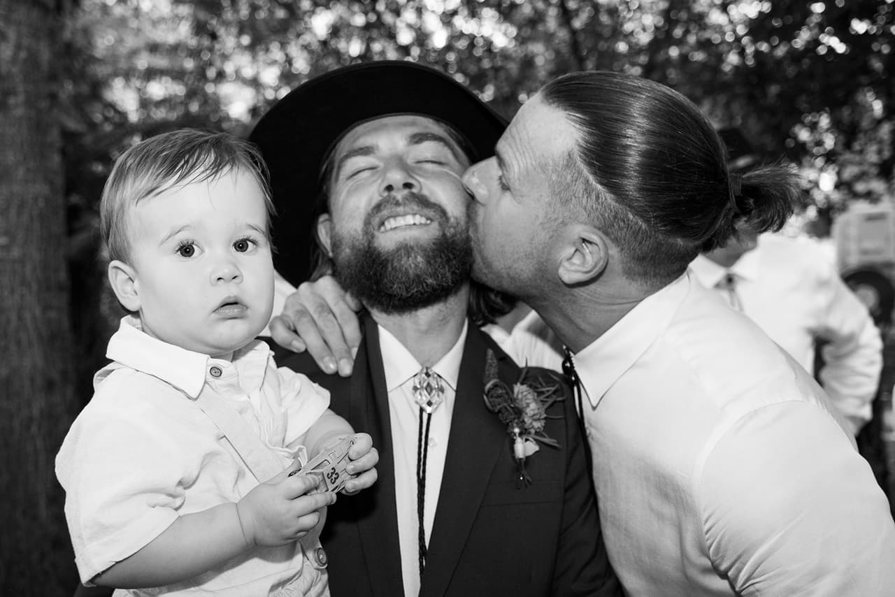 Black and white photo of a groomsman kissing Todd on the cheek while Todd holds a baby boy