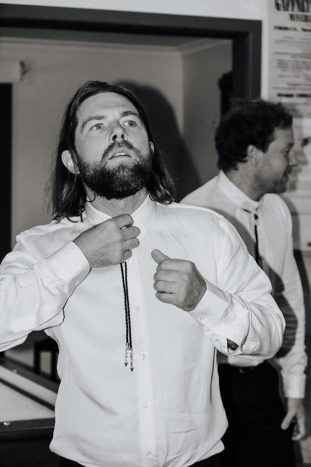 Black and white photo of Todd doing up his bolo tie in the pub, a groomsman beside him