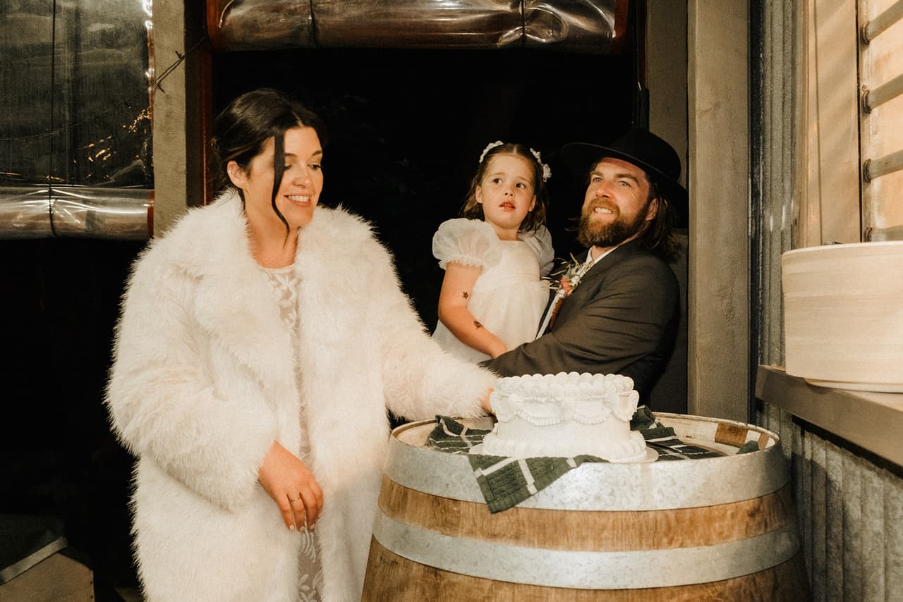 Lana in a white faux fur coat cutting the cake on a wine barrel with Todd and a flower girl