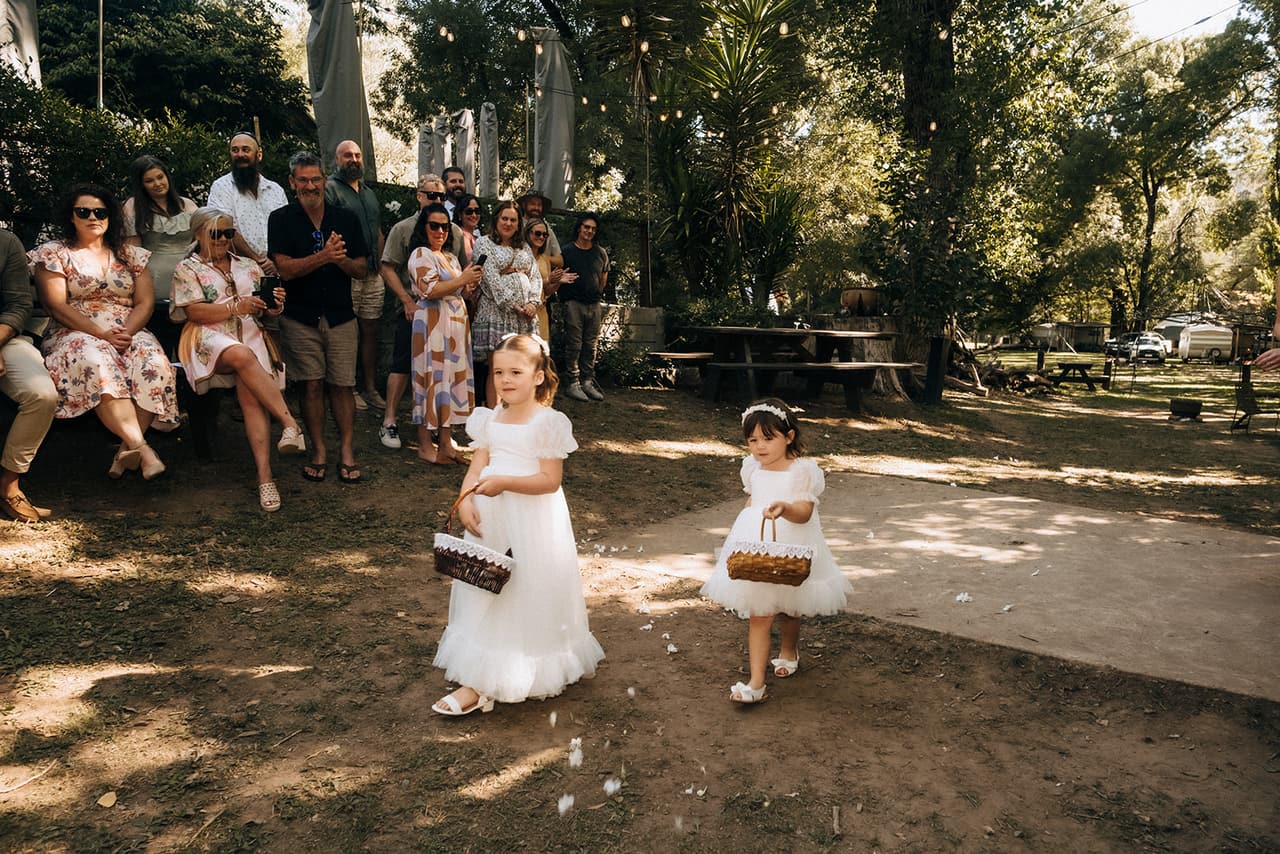 Two flower girls in white walking down the aisle with their baskets as the guests watch