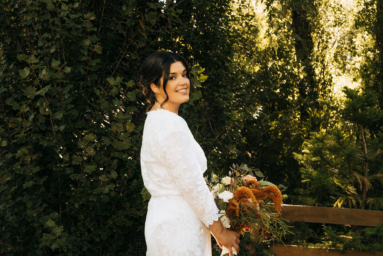 Lana with her bouquet, looking back over her shoulder beside a timber fence in the trees