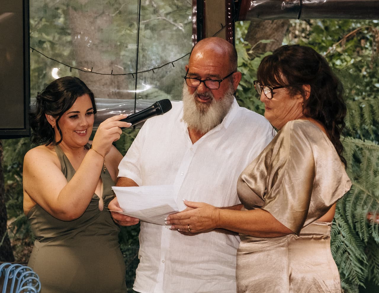 Lana’s dad reading his speech beside a woman in champagne satin while a bridesmaid holds the microphone