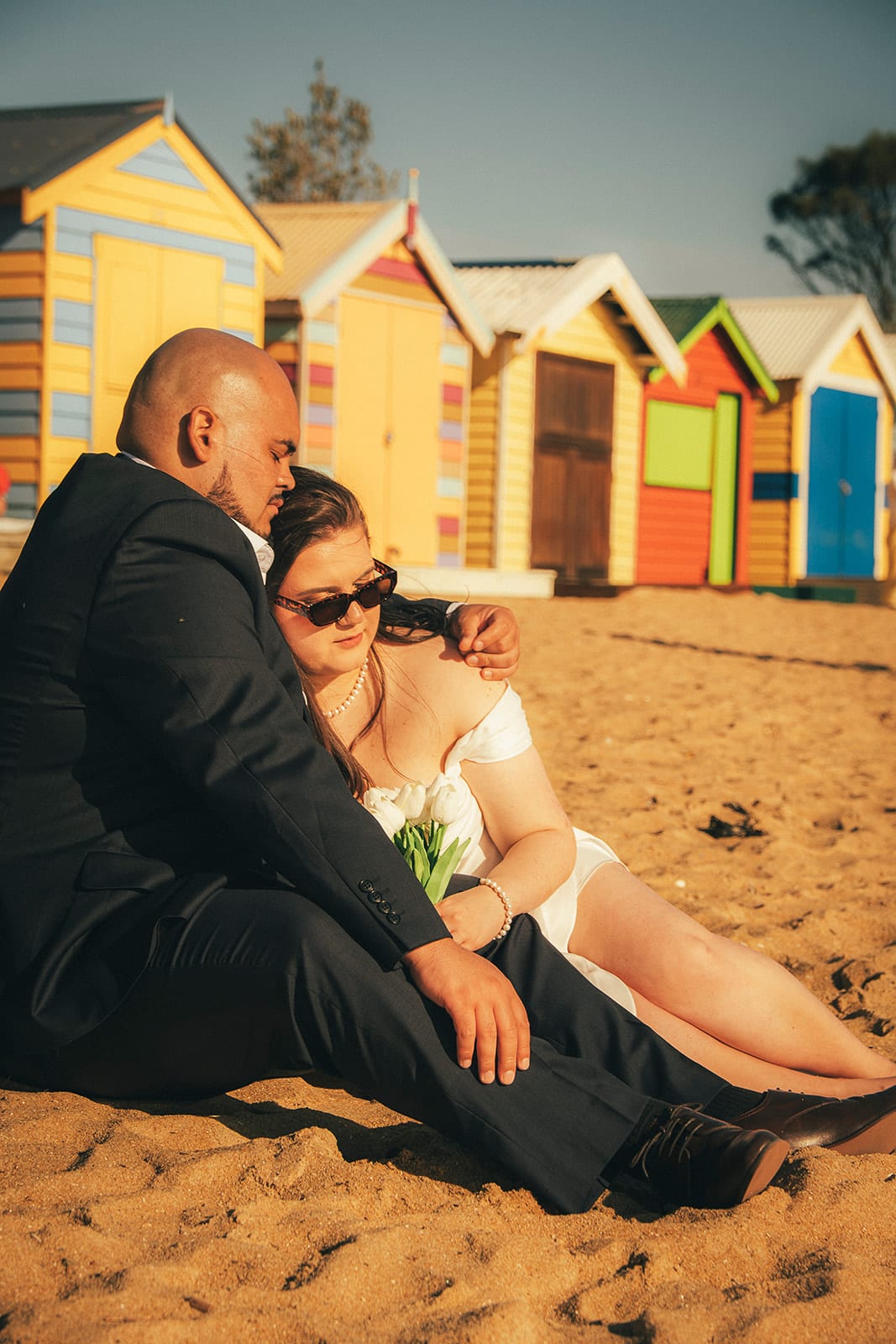 Martina and Diego sitting on the sand in front of the striped bathing boxes, sunglasses on