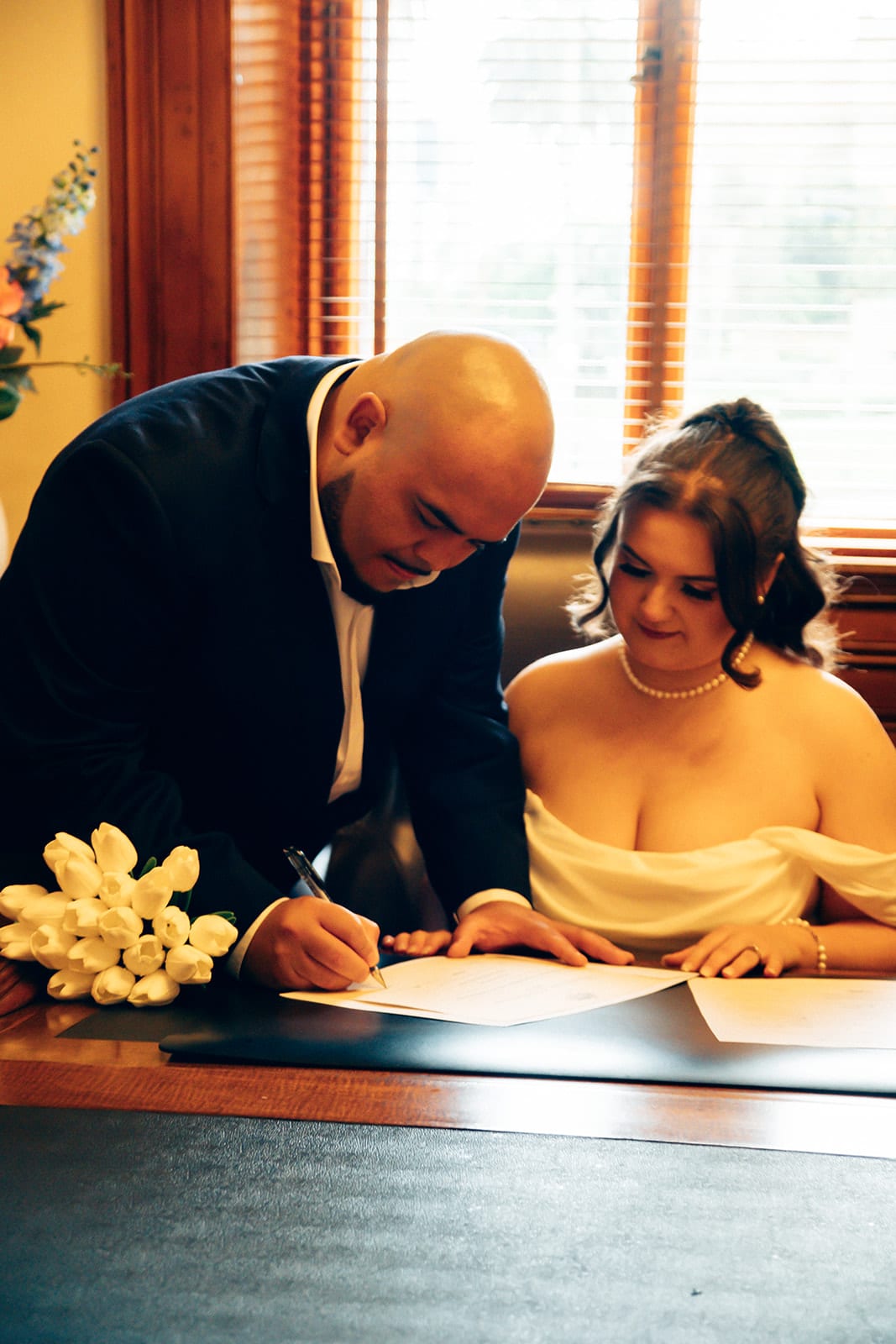 Diego signing at the registry desk while Martina sits beside him, her tulips on the desk