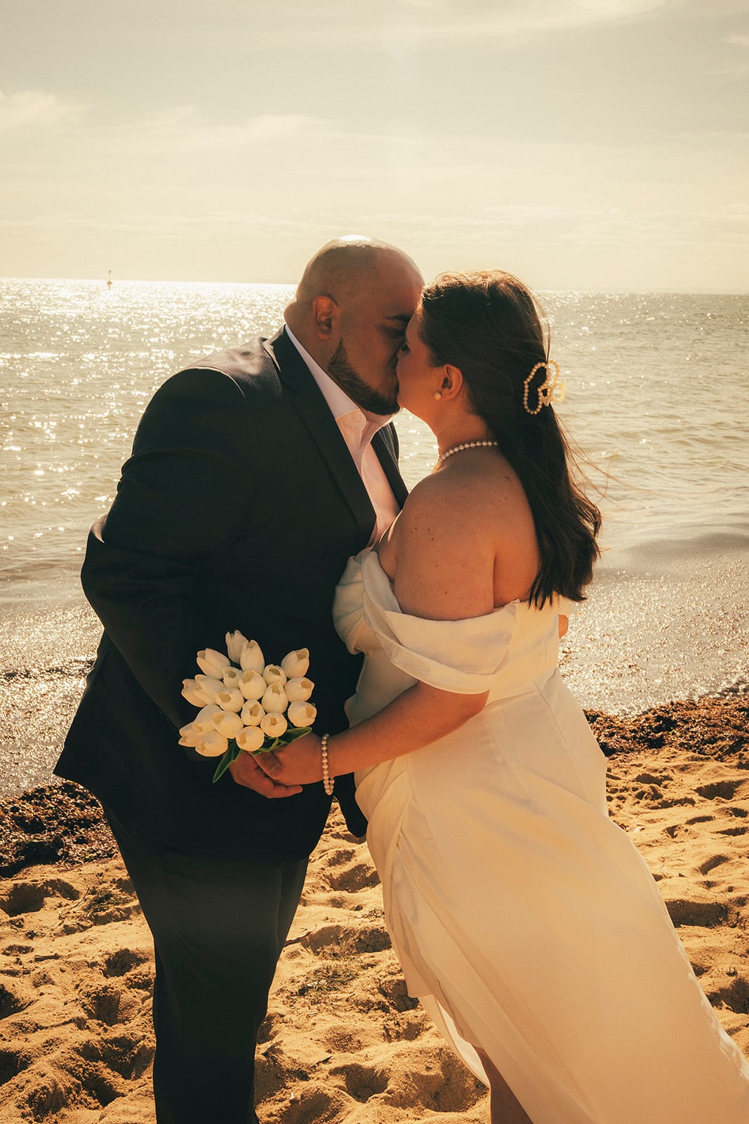 Martina and Diego kissing on the sand, her tulips in his hand and the bay glittering behind them