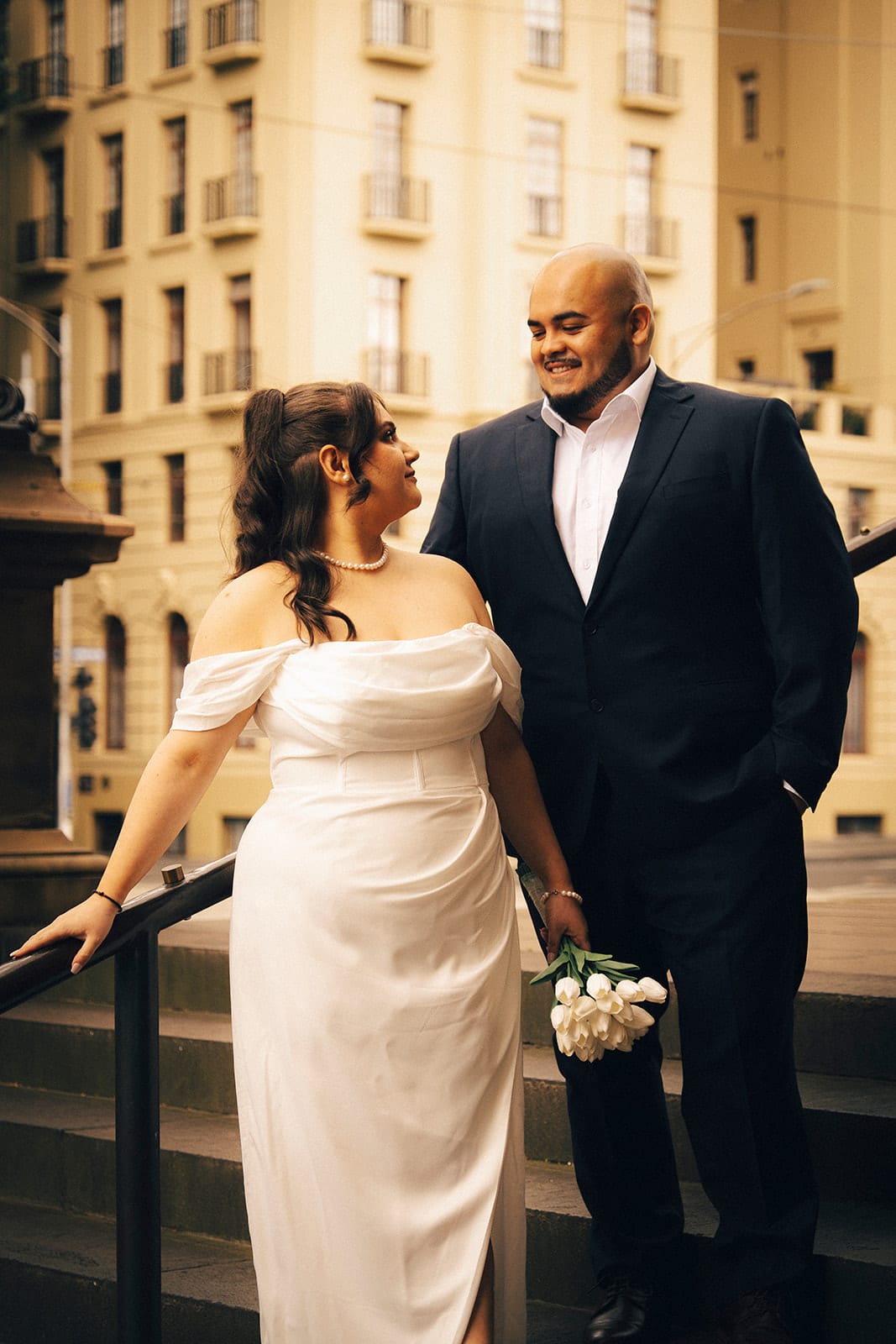 Martina laughing up at Diego as she holds the railing of the stone steps, a tall cream building behind them