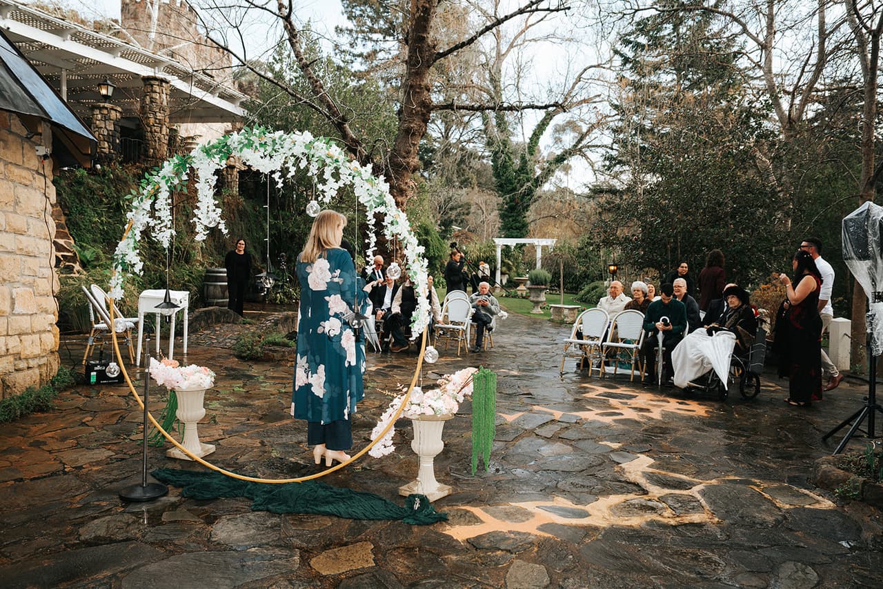 The ceremony set beside the little stone chapel, a white floral arch over the wet stone and guests seated beyond