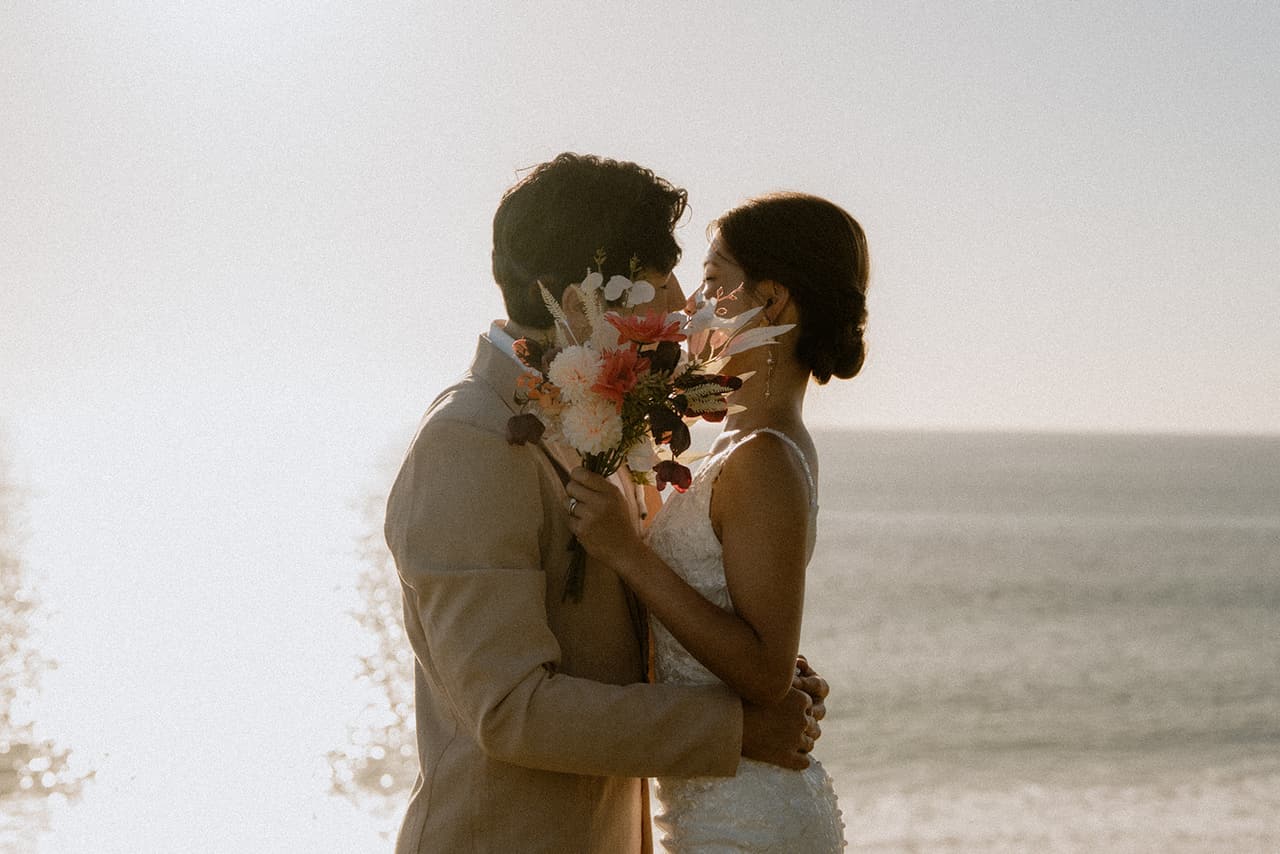 Sayuri and Shuto kissing behind her bouquet in hazy afternoon light above the sea