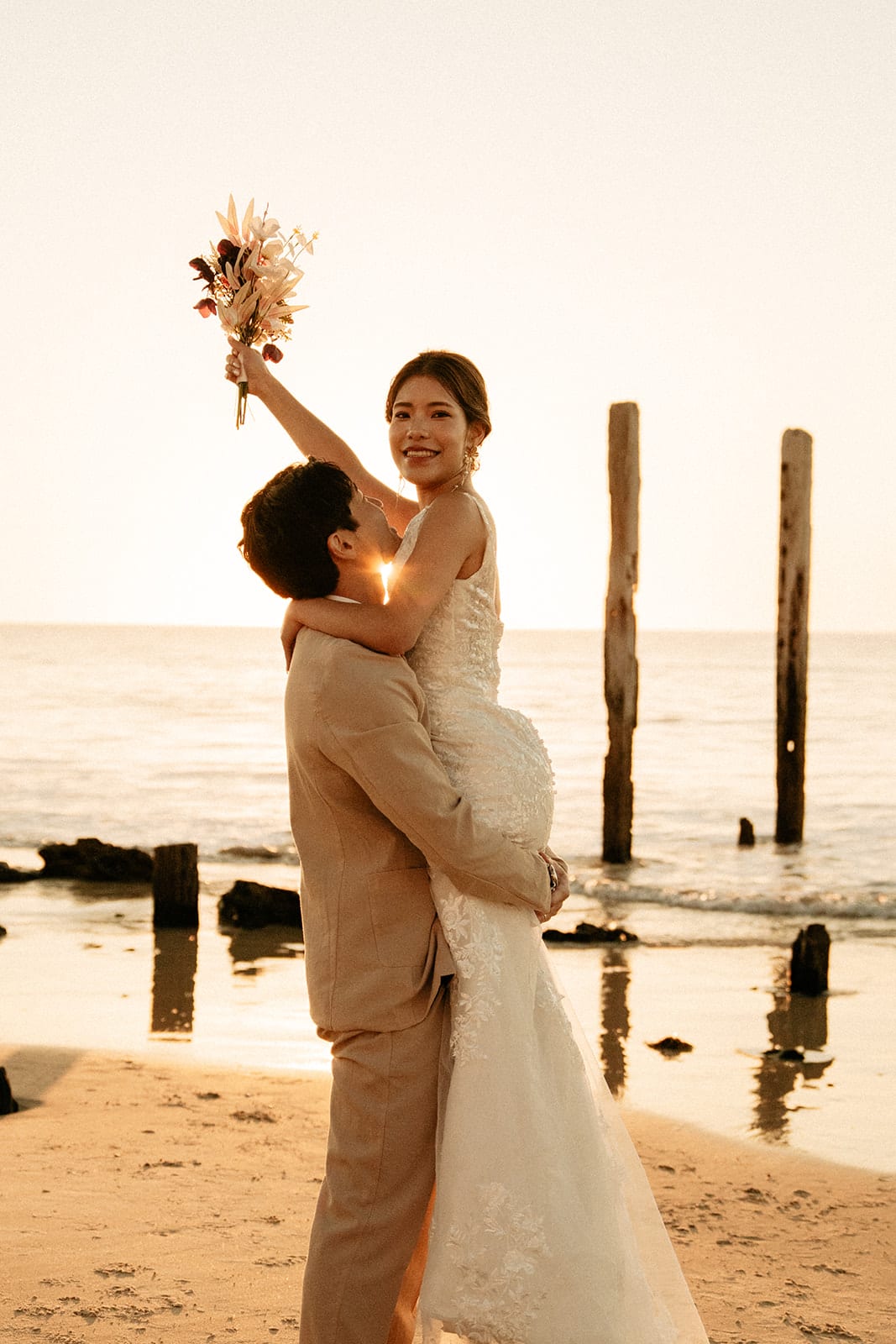 Sayuri raising her bouquet to the sky as Shuto lifts her beside the jetty posts at sunset