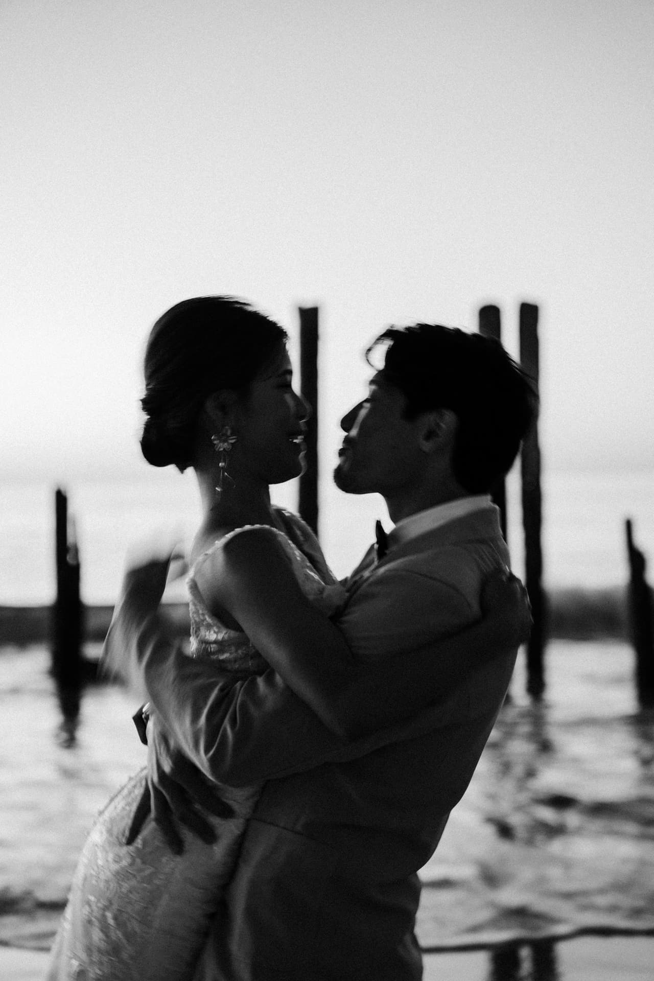Black and white photo of Sayuri and Shuto embracing among the old jetty posts