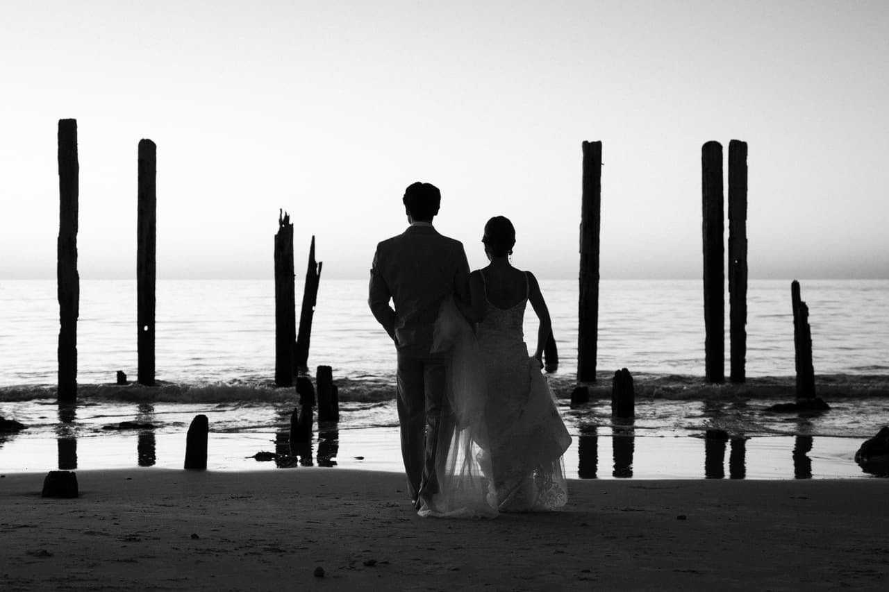 Black and white photo of Sayuri and Shuto walking towards the old jetty posts at the water’s edge