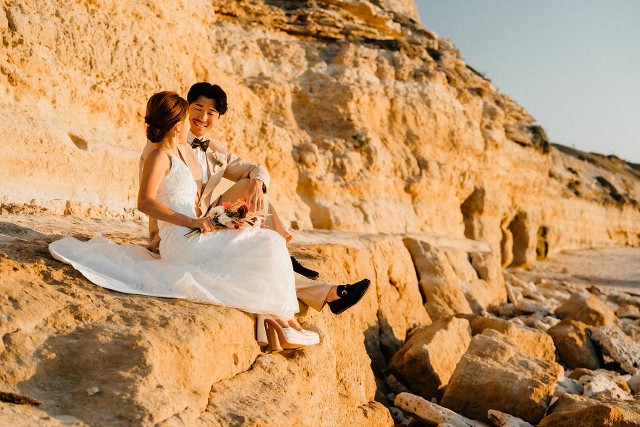 Sayuri and Shuto sitting together on a sandstone ledge at the foot of the cliffs, small caves in the cliff face behind them