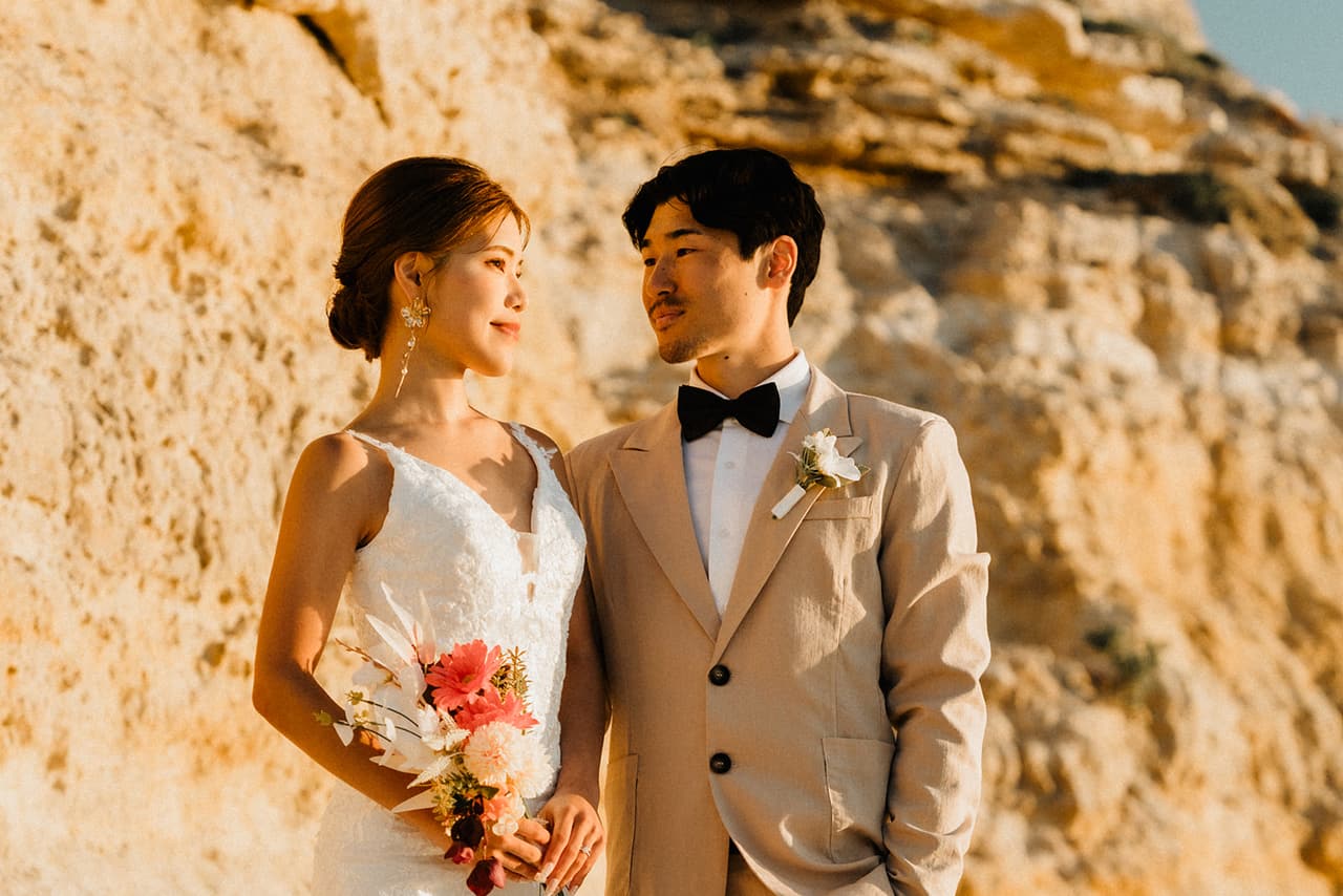 Sayuri and Shuto standing close against the golden sandstone cliff, smiling at each other
