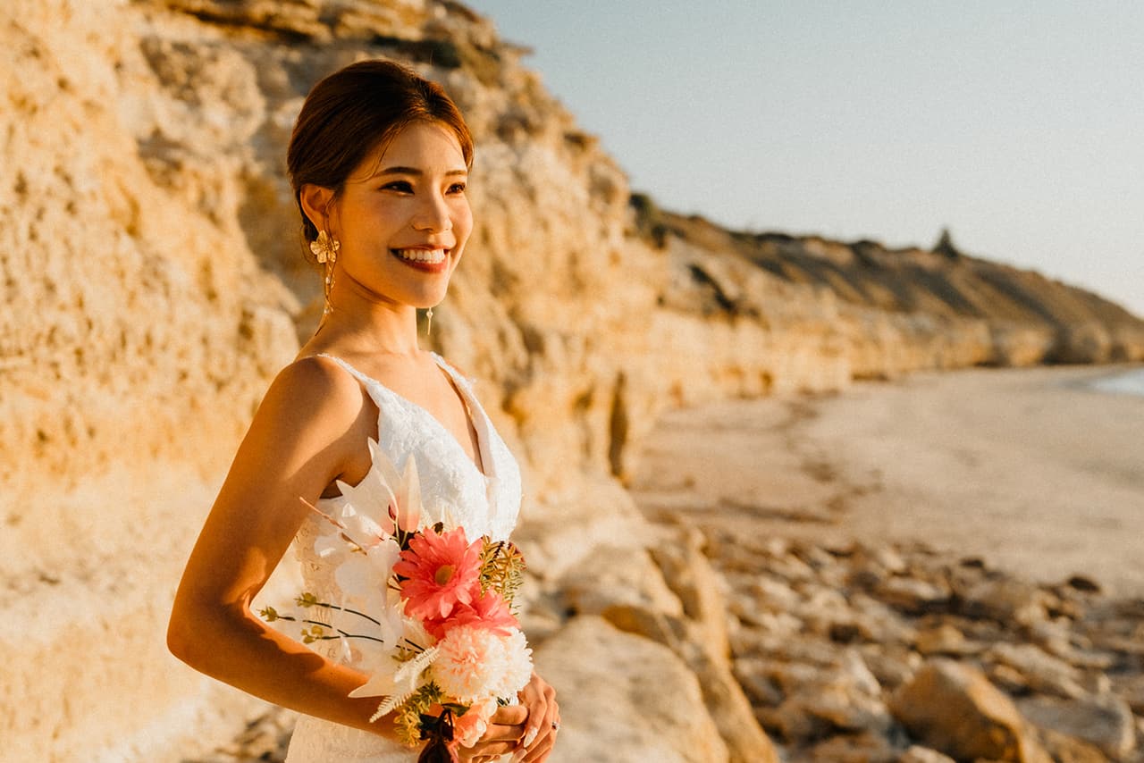 Sayuri laughing with her bouquet against the golden cliffs, the beach stretching away behind her