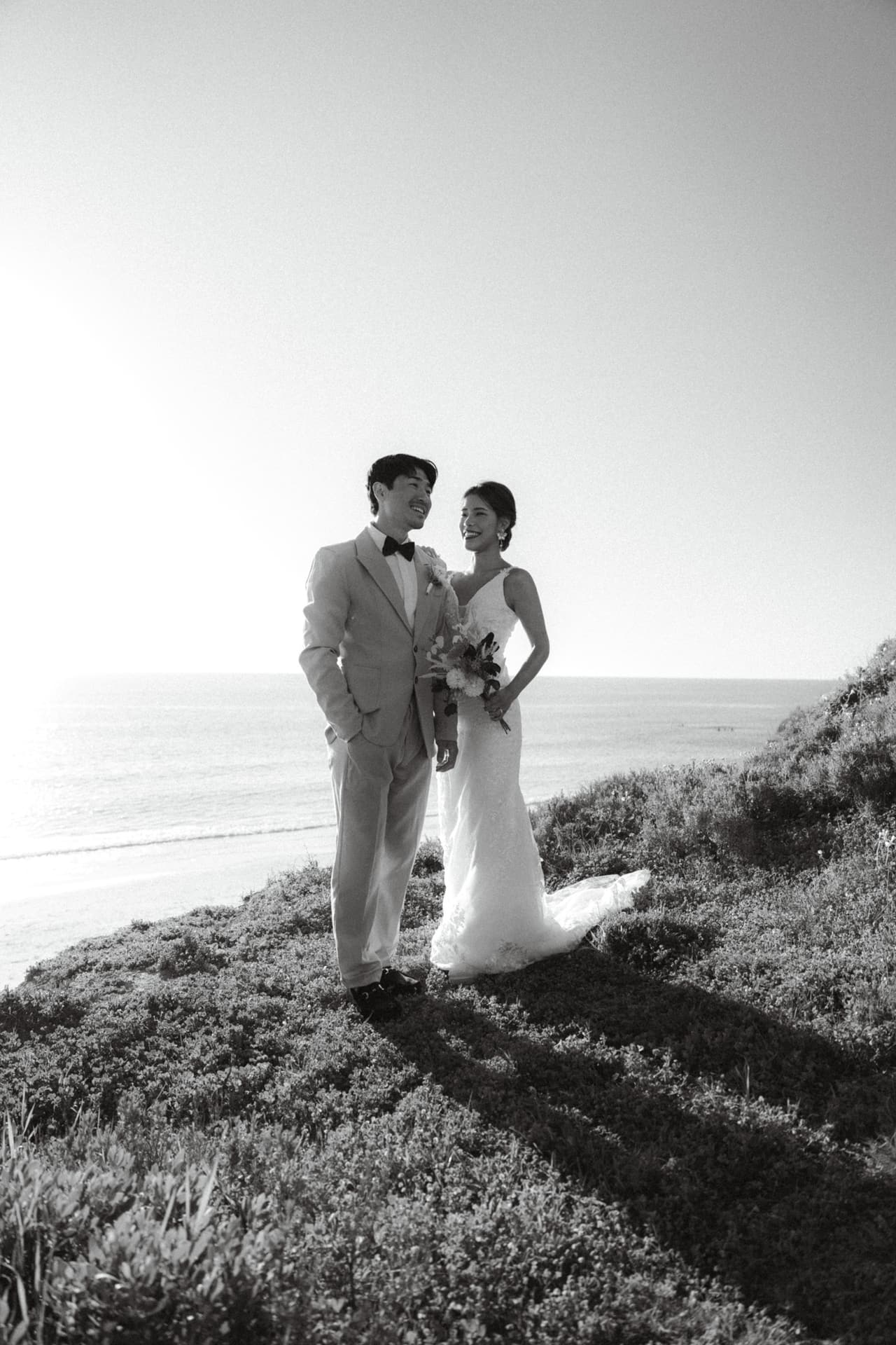 Sayuri and Shuto standing on a grassy cliff above the sea, the sun behind them