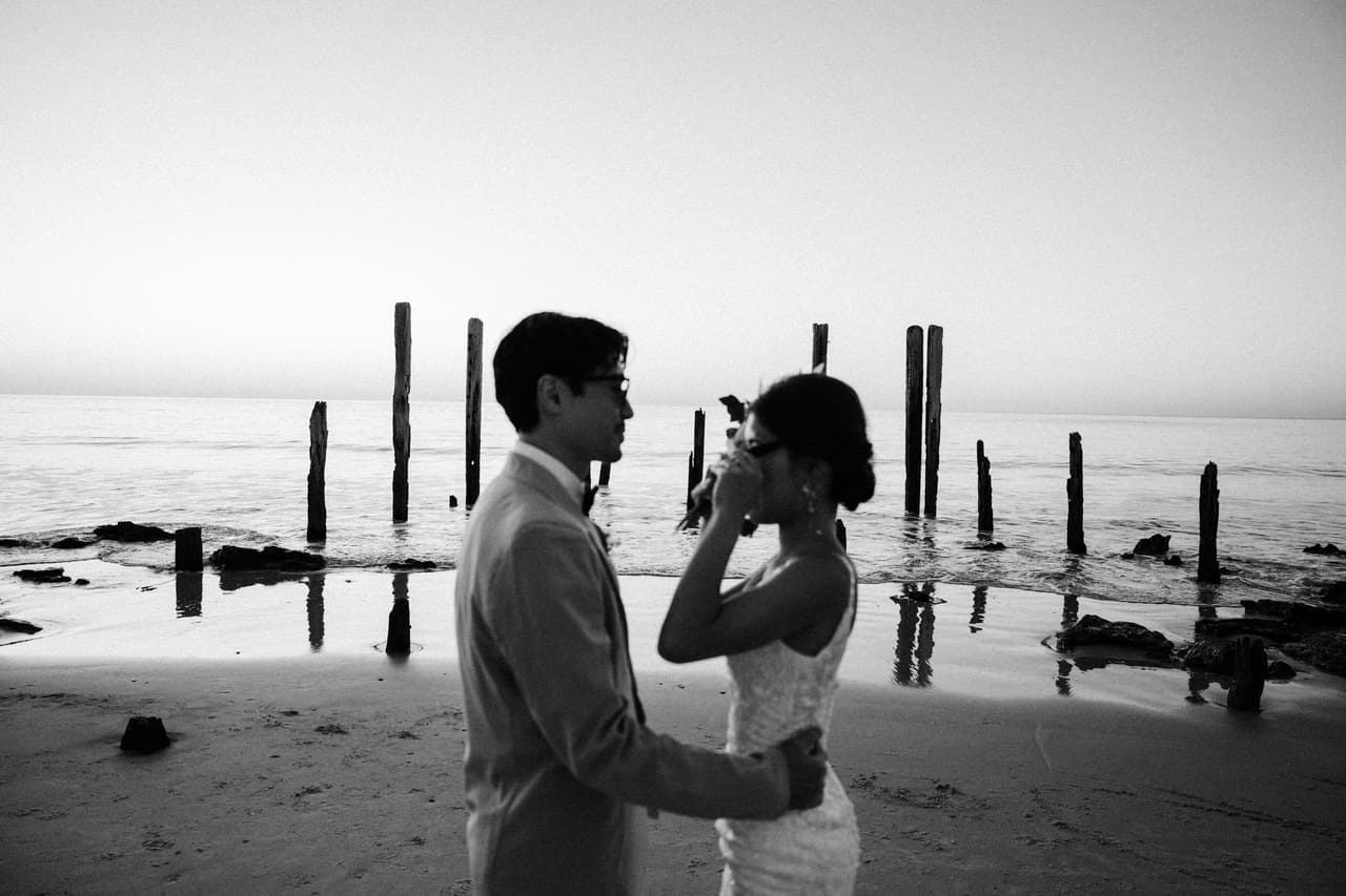 Black and white photo of Sayuri and Shuto facing each other on the beach beside the old jetty posts