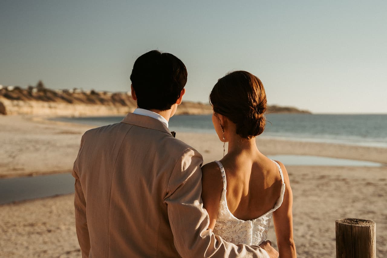 Sayuri and Shuto seen from behind, looking at each other on the beach beside a weathered timber post