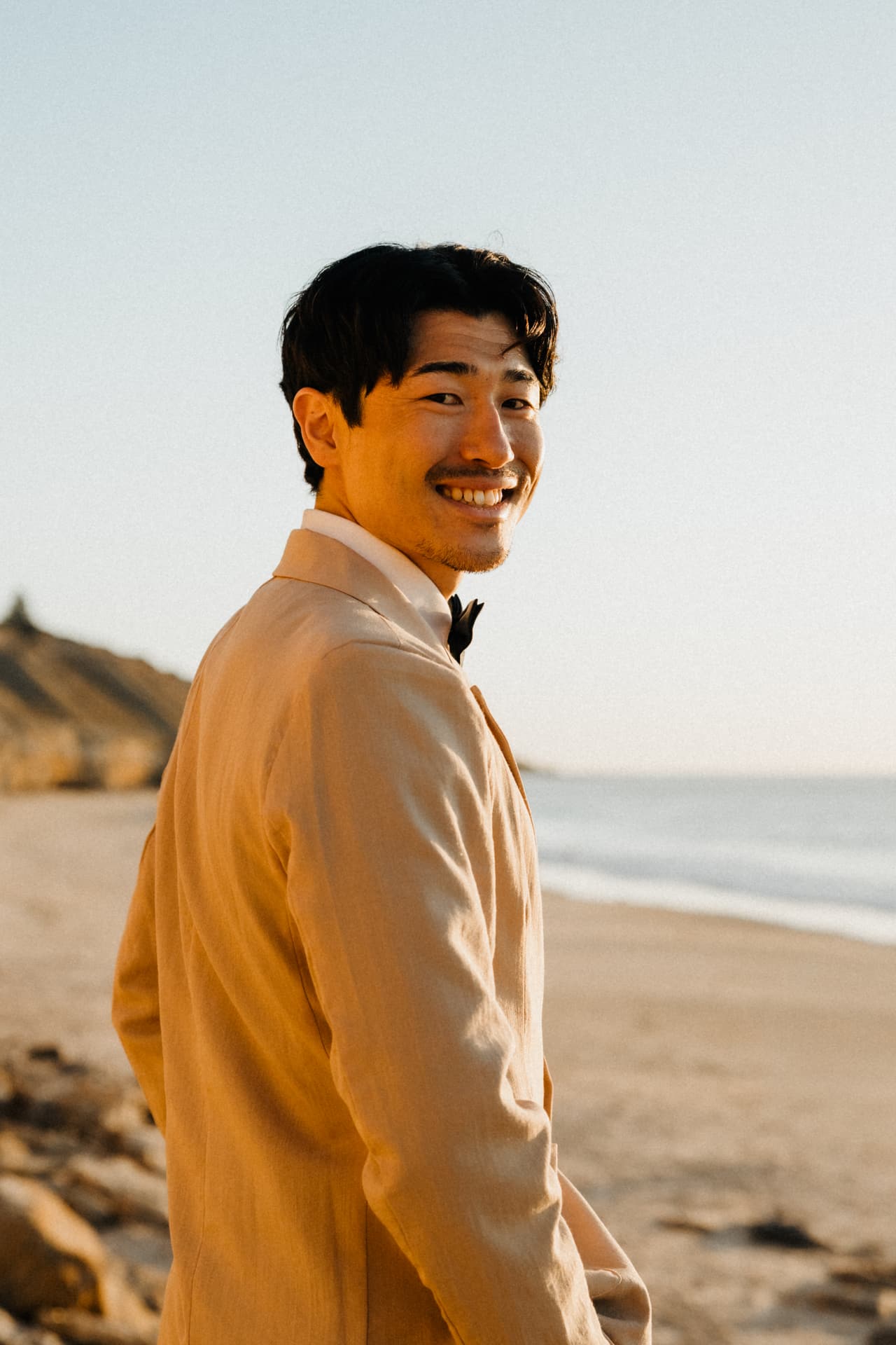 Shuto smiling over his shoulder in his beige suit, the beach, the cliffs and the sea behind him