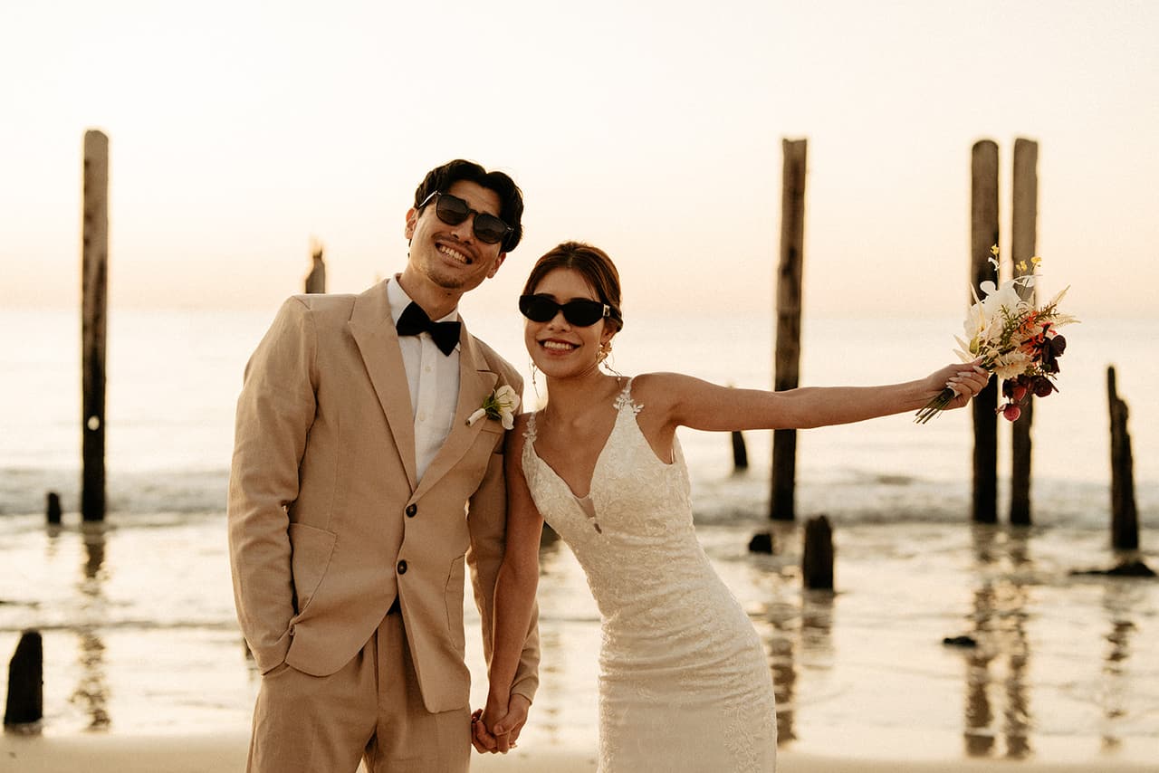 Shuto and Sayuri in sunglasses among the jetty posts, Sayuri stretching out her arm with the bouquet
