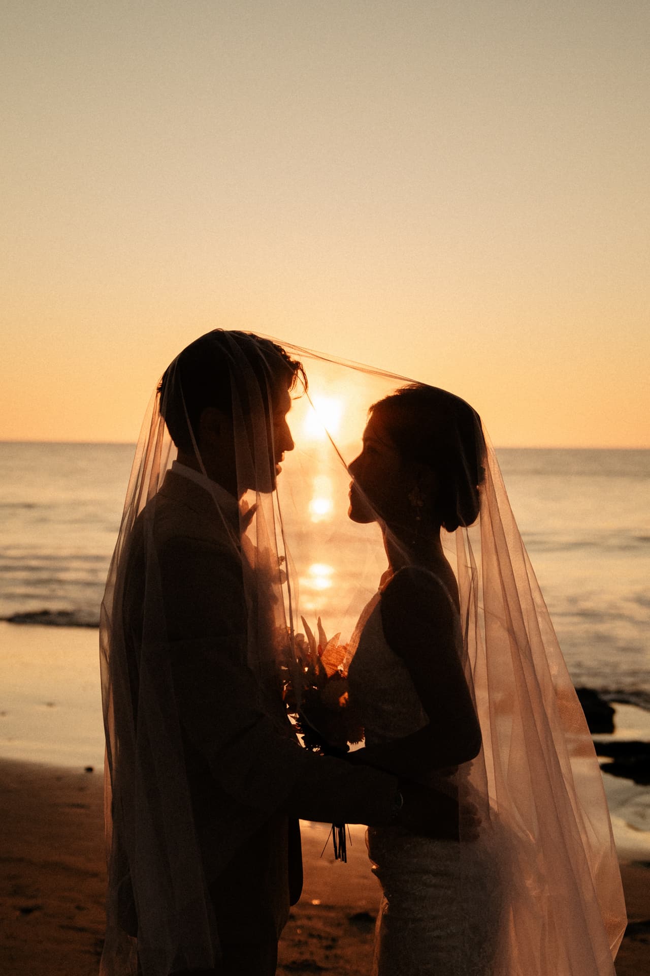 Sayuri and Shuto silhouetted beneath her veil on the beach at sunset
