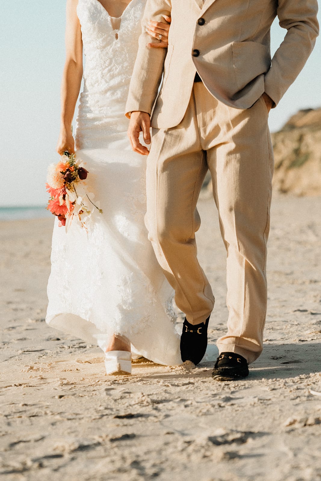 Sayuri and Shuto walking arm in arm across the sand, her bouquet at her side, seen from the waist down