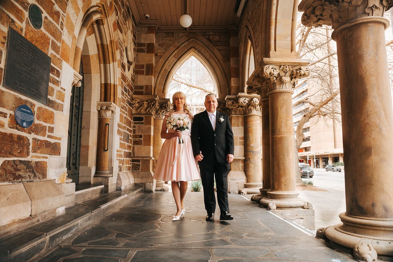 Terrie and Jon walking hand in hand under a sandstone arcade of Gothic arches and carved columns