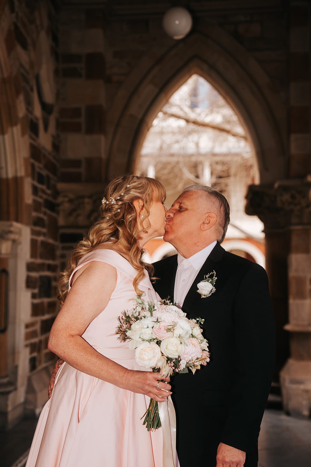 Jon kissing Terrie under a pointed sandstone arch, her bouquet of pale pink and white roses in her hand