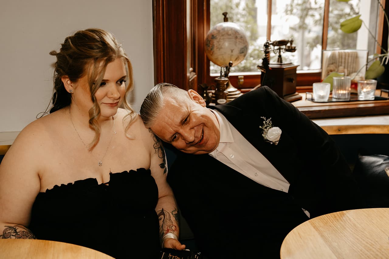 Jon resting his head on the shoulder of a young woman in black as they laugh together in a booth