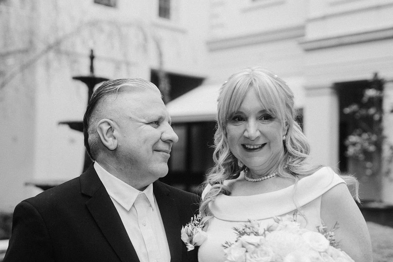 Black and white photo of Jon and Terrie smiling side by side in the courtyard, the fountain behind them