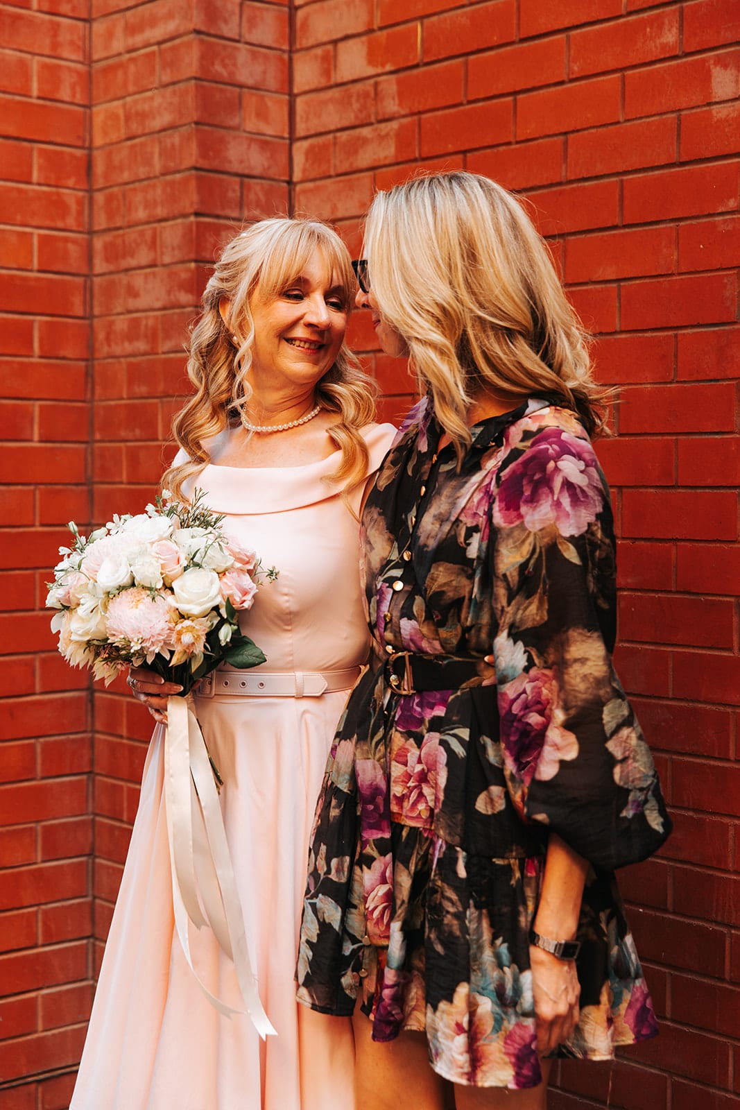 Terrie and a friend in a dark floral dress leaning their heads together in front of a red brick wall