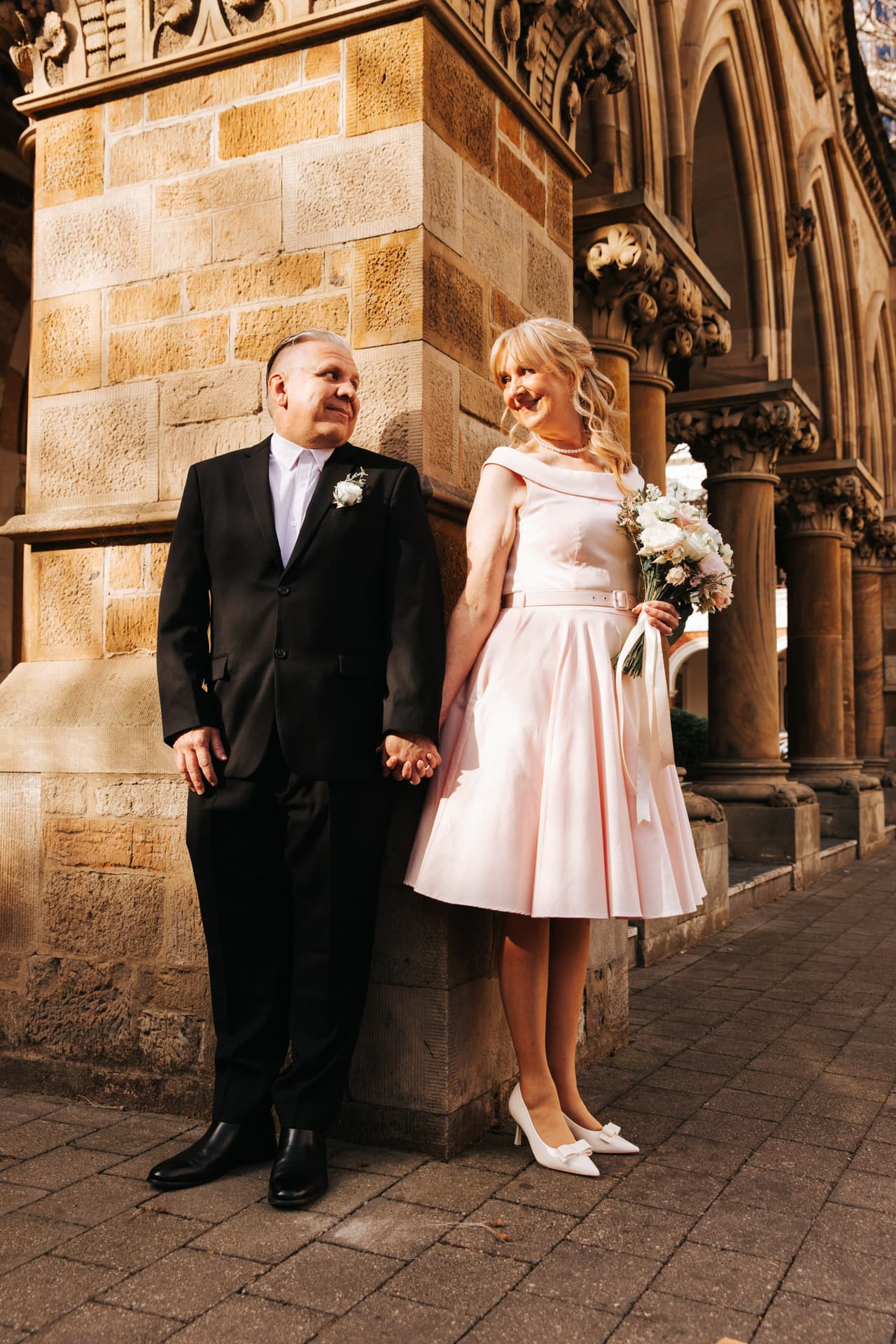 Terrie in a pink tea-length dress and Jon holding hands beside sandstone Gothic arches