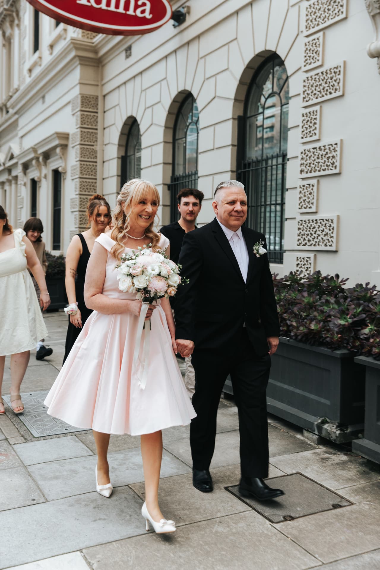 Terrie and Jon walking along a city footpath past a white building with arched windows, their guests following behind