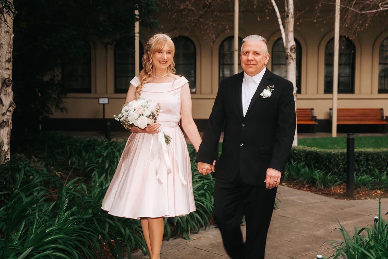Terrie and Jon walking hand in hand through a green courtyard beside a sandstone building